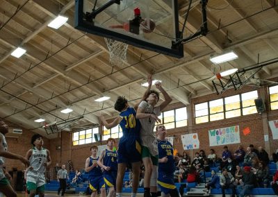A photograph of an active WSBC basketball game in the ASDB gymnasium. On court are the competing Utah Eagles and Washington Terrier boys teams. In the photo, a Terrier player attempts a layup over the arm of a threatening defender.