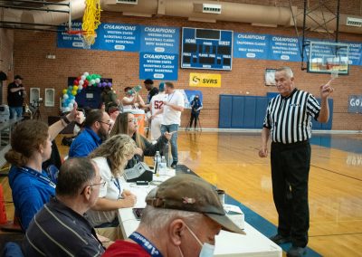A photograph of an active WSBC basketball game in the ASDB gymnasium. Beside the court is a long, white table where the game scorekeepers are seated. Standing in front of them is the match referee who is gesturing with his left hand.
