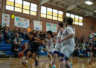 A photograph of an active WSBC basketball game in the ASDB gymnasium. On court are the competing Arizona Sentinel and Oregon Panther boys teams. In the photo, a Sentinel Player is dribbling towards the basket with three Oregon defenders in front of him.