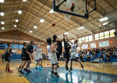 A photograph of an active WSBC basketball game in the ASDB gymnasium. On court are the competing Arizona Sentinel and Oregon Panther boys teams. In the photo, a Sentinel player is attempting a shot from underneath the basket.