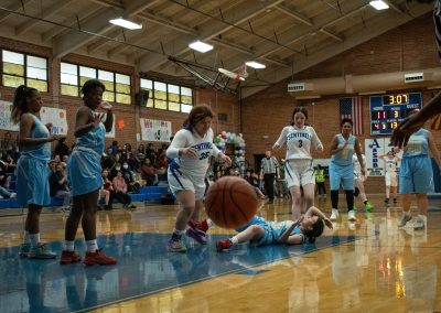 A photograph of an active WSBC basketball game in the ASDB gymnasium. On court are the competing Phoenix Roadrunner and Arizona Sentinel girls teams. In the photo, a Roadrunner player has fallen over and lost control of the basketball.