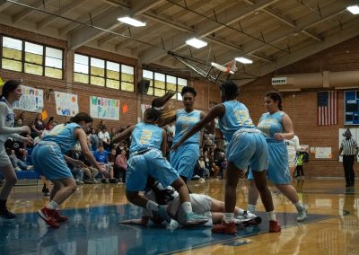 A photograph of an active WSBC basketball game in the ASDB gymnasium. On court are the competing Phoenix Roadrunner and Arizona Sentinel girls teams. In the photo, a Sentinel player falls over after being stuffed by the entire Roadrunner team.