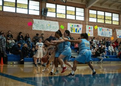 A photograph of an active WSBC basketball game in the ASDB gymnasium. On court are the competing Phoenix Roadrunner and Arizona Sentinel girls teams. In the photo, a driving Sentinel player is being covered by three separate Roadrunner defenders.
