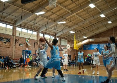 A photograph of an active WSBC basketball game in the ASDB gymnasium. On court are the competing Phoenix Roadrunners and Arizona Sentinel girls teams. In the photo, a Sentinel player is preparing to take a shot from underneath the basket.