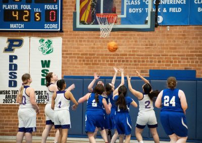 A photograph of an active WSBC basketball game in the ASDB gymnasium. On court are the competing Utah Eagle and Oregon Panther girls teams. In the photo, a crowd of Eagle players prepare to secure a basketball rebound before any of the Panther players can.