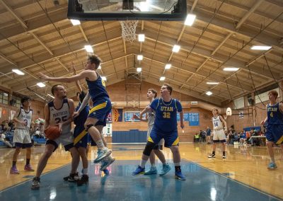 A photograph of an active WSBC basketball game in the ASDB gymnasium. On court are the competing Utah Eagle and Oregon Panther boys teams. In the photo, a leaping Eagle defender prevents a Panther player from attempting a shot from the paint.