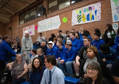 A photo of a crowd gathered in the bleachers of the ASDB gymnasium. Prominently featured is the entire Utah Eagles boys team. Behind them, running the length of the gym's brick wall, are multiple posters encouraging the teams participating in WSBC.