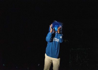 A photograph of Coach Gerald Brown standing in the center of a bright spotlight. He is wearing a disguise which features a blue wig and a blue face mask. He is pictured signing frantically.