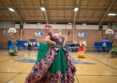 A photograph of multiple individuals dancing on the basketball court in the ASDB gymnasium. Each is wearing an ornate dress with flowery designs. They are members of the Arizona Folklórico Dance Company, a WSBC sponsor.