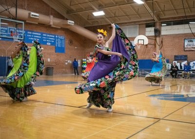 A photograph of multiple individuals dancing on the basketball court in the ASDB gymnasium. Each is wearing an ornate dress with flowery designs. They are members of the Arizona Folklórico Dance Company, a WSBC sponsor.