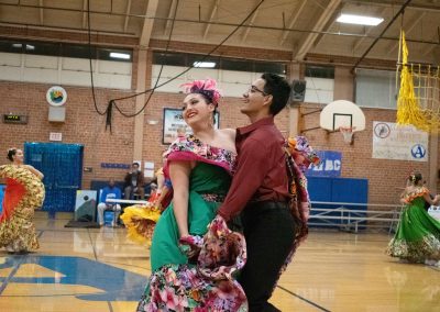 A photograph of two individuals dancing on the basketball court in the ASDB gymnasium. One of them is wearing an ornate flowery dress of pink, green and white. The other is wearing a maroon button-up shirt and black slacks. They are both members of the Arizona Folklórico Dance Company, a WSBC sponsor.