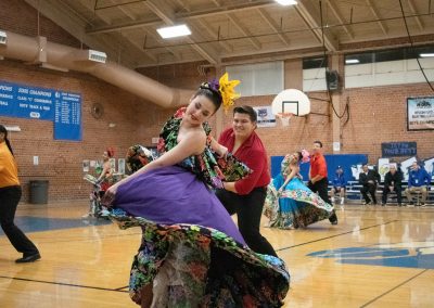 A photograph of two individuals dancing on the basketball court in the ASDB gymnasium. One of them is wearing an ornate flowery dress of purple, green and black. The other is wearing a red button-up shirt and black slacks. They are both members of the Arizona Folklórico Dance Company, a WSBC sponsor.