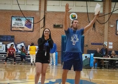 A photograph of two individuals standing at the far edge of the basketball court in the ASDB gymnasium. They are raising their arms and clapping to an audience not pictured within the frame.