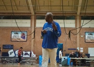 A photograph of coach Gerald Brown standing at the far edge of the basketball court in the ASDB gymnasium. He is pictured signing the number four.