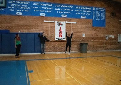 A photograph of three individuals standing in the ASDB gymnasium. They are unveiling banners which honor the teams participating in WSBC. After having revealed the Idaho Raptor's team banner, they are showing raising their arms in celebration.