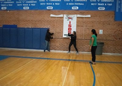 A photograph of three individuals standing in the ASDB gymnasium. They are unveiling banners which honor the teams participating in WSBC. One of them is pictured tugging on a rope in order to reveal the next banner.