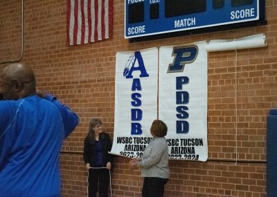 A photograph of ASDB Superintendent Annette Reichman and Assistant Superintendent Shaina Cooper standing against a brick wall in the ASDB gymnasium. The two are pictured smiling and communicating. Behind them are large, hung-up WSBC banners honoring ASDB and PDSD, respectively.