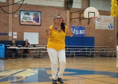 A photograph of an individual in a yellow shirt standing at half court in the ASDB gymnasium. They are passionately signing to an audience not pictured within the frame.