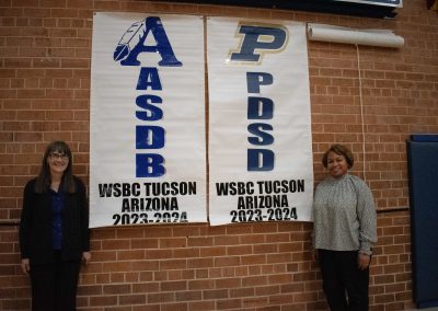 A photograph of ASDB Superintendent Annette Reichman and Assistant Superintendent Shaina Cooper standing against a brick wall in the ASDB gymnasium. Behind them are large, hung-up WSBC banners honoring ASDB and PDSD, respectively.