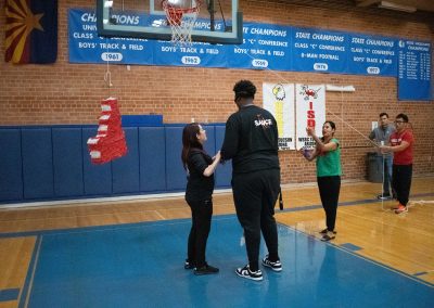 A photograph of a blindfolded individual preparing to strike a piñata in the ASDB gymnasium. The piñata is colored red and white, and is printed with the Idaho Raptor's athletic logo. It is also crafted to look like the state of Idaho.