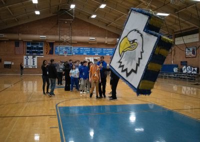 A photograph of a hung piñata in the ASDB gymnasium. The piñata is colored blue and white, and is printed with the Utah Eagle's athletic logo. Behind the decoration is a line of students waiting for their turn to take a swing.