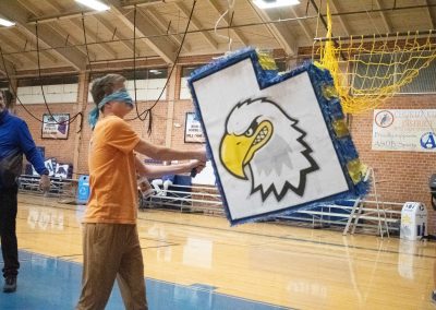 A photograph of a blindfolded student striking a piñata in the ASDB gymnasium. The piñata is colored blue and white, and is printed with the Utah Eagle's athletic logo. It is also crafted to look like the state of Utah.