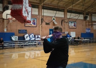 A photograph of a blindfolded student striking a piñata in the ASDB gymnasium. The piñata is colored red and white, and is printed with the Idaho Raptor's athletic logo. It is also crafted to look like the state of Idaho.