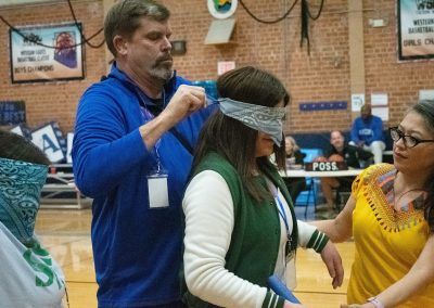 A photograph of a student standing at the far side of the ASDB gymnasium. The student is standing patiently while a separate individual ties a blindfold over their eyes.
