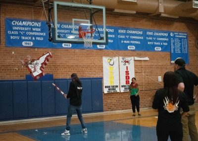 A photograph of a student striking a piñata in the ASDB gymnasium. The piñata is colored red and white, and is printed with the Idaho Raptor's athletic logo. The floor below is covered with candy and debris from the decoration.