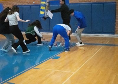 A photograph of the ASDB gymnasium floor. Spread across the floor are remnants of a blue and yellow piñata as well as various bits of candy. Students are pictured scrambling to collect the candy.