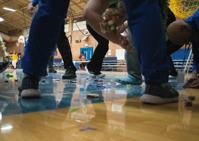 A close-up photograph of the ASDB gymnasium floor. Spread across the floor are remnants of a blue and yellow piñata, as well as various bits of candy. Students are pictured scrambling to collect the candy.