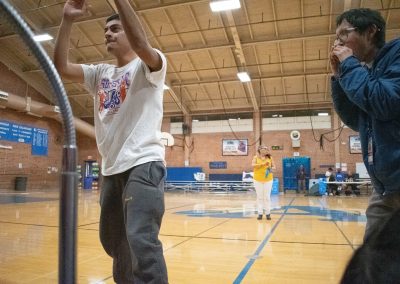 A photograph of two students standing at the edge of the basketball court in the ASDB gymnasium. One student is pictured clapping their hands above their head. The other students is cupping their hands over their mouth and yelling.