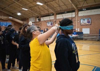 A photograph of a line of Phoenix Roadrunner students standing at the far side of the ASDB gymnasium. At the front of the line, one student stands patiently while a separate individual ties a blindfold over their eyes.