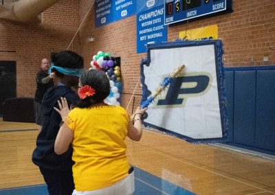 A photograph of a student preparing to strike a piñata in the ASDB gymnasium. The piñata is colored blue and white, and is printed with the Phoenix Roadrunner's athletic logo. Another individual is standing next to the student and helping them to locate the piñata with a bat.