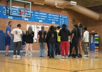 A photograph of a crowd standing at the far side of the ASDB gymnasium. They are watching as a large piñata is being hoisted up into the air. Just below the decoration, a blindfolded student is preparing to take a swing.