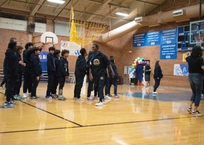 A photograph of a student walking across the basketball court in the ASDB gymnasium. The student is smiling widely after attempting a few swings at a hung piñata. Behind the student, a line of additional students wait for their turn to take a swing.