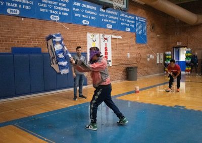 A photograph of a student striking a piñata in the ASDB gymnasium. The piñata is colored blue and white, and is crafted to look like the state of Arizona. Printed on the outside of the piñata is the Arizona Sentinel's athletic logo.