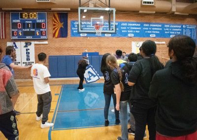 A photograph of a student striking a piñata in the ASDB gymnasium. The piñata is colored blue and white, and is printed with the Arizona Sentinel's athletic logo. Behind them is a line of students waiting for their turn to swing.