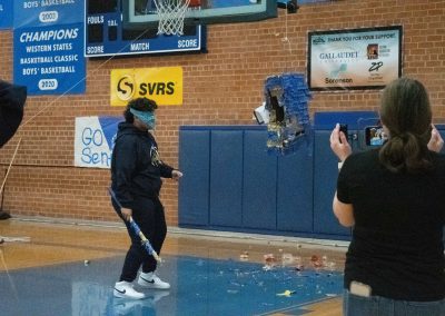 A photograph of a student standing in the ASDB gymnasium. The student is striking a blue piñata with a long stick. The floor below is covered with candy and piñata debris.
