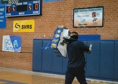 A photograph of a student standing in the ASDB gymnasium. The student is striking a blue piñata with a long stick. The piñata is colored blue and white, and is printed with the Phoenix Roadrunner's athletic logo.