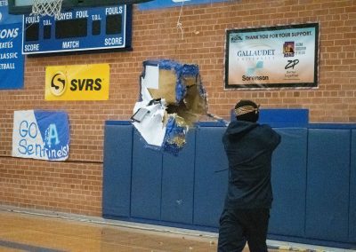 A photograph of a student standing in the ASDB gymnasium. The student is striking a blue piñata with a long stick. The piñata is colored blue, white and gold, and is crafted to look like the state of Arizona.
