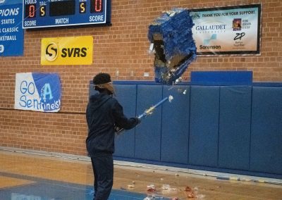 A photograph of a student standing in the ASDB gymnasium. They are striking a blue piñata with a long stick. The floor below is covered with candy and piñata debris.
