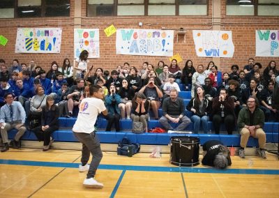 A photograph of a crowd gathered in the bleachers of the ASDB gymnasium. Standing in front of them is a student beating on a large hand drum. Members of the audience are pictured with their hands cupped around their mouths, gleefully responding back to the student with yells.
