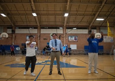 A photograph of ASDB Principal Paul Saunders, Coach Gerald Brown, and a student, standing in the center of the basketball court in the ASDB gymnasium. They are signing to an audience not pictured within the frame.